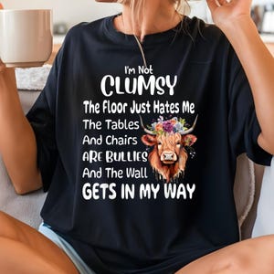May include: Black t-shirt with a white graphic of a Highland cow with flowers in its horns. The text "I'm Not Clumsy The Floor Just Hates Me The Tables And Chairs Are Bullies And The Wall Gets In My Way" is printed on the shirt.