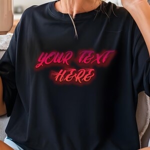May include: Black crew neck t-shirt with the text "YOUR TEXT HERE" in a gradient pink and red font. The text has a neon-like glow. The shirt is a solid black color.