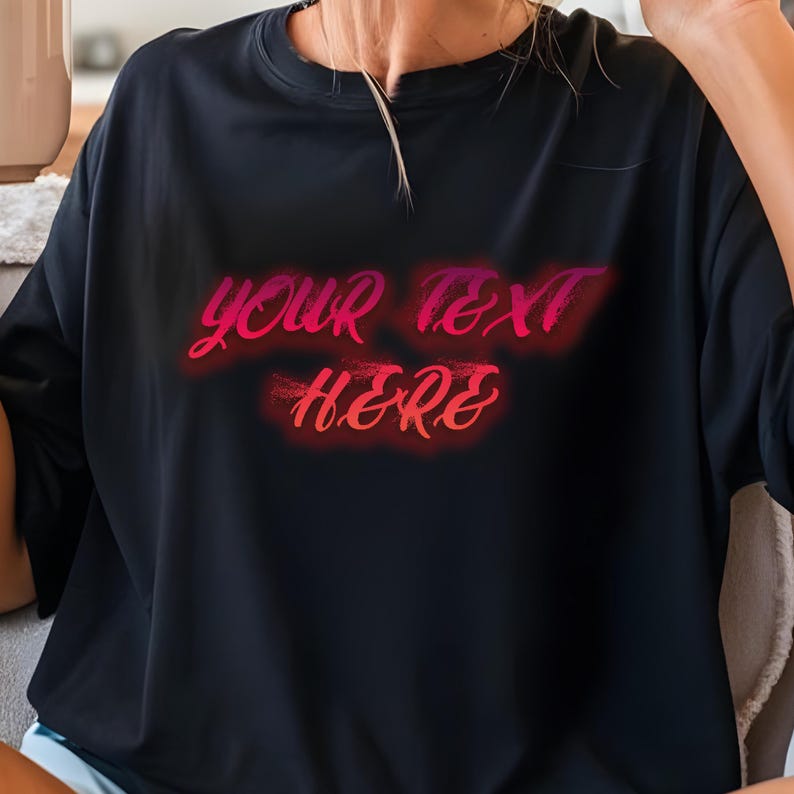 May include: Black crew neck t-shirt with the text "YOUR TEXT HERE" in a gradient pink and red font. The text has a neon-like glow. The shirt is a solid black color.