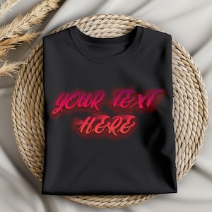 May include: A black t-shirt with the words "YOUR TEXT HERE" in a gradient red and pink font. The shirt is folded neatly and rests on a woven, circular placemat. Dried wheat stalks are visible in the upper left corner.