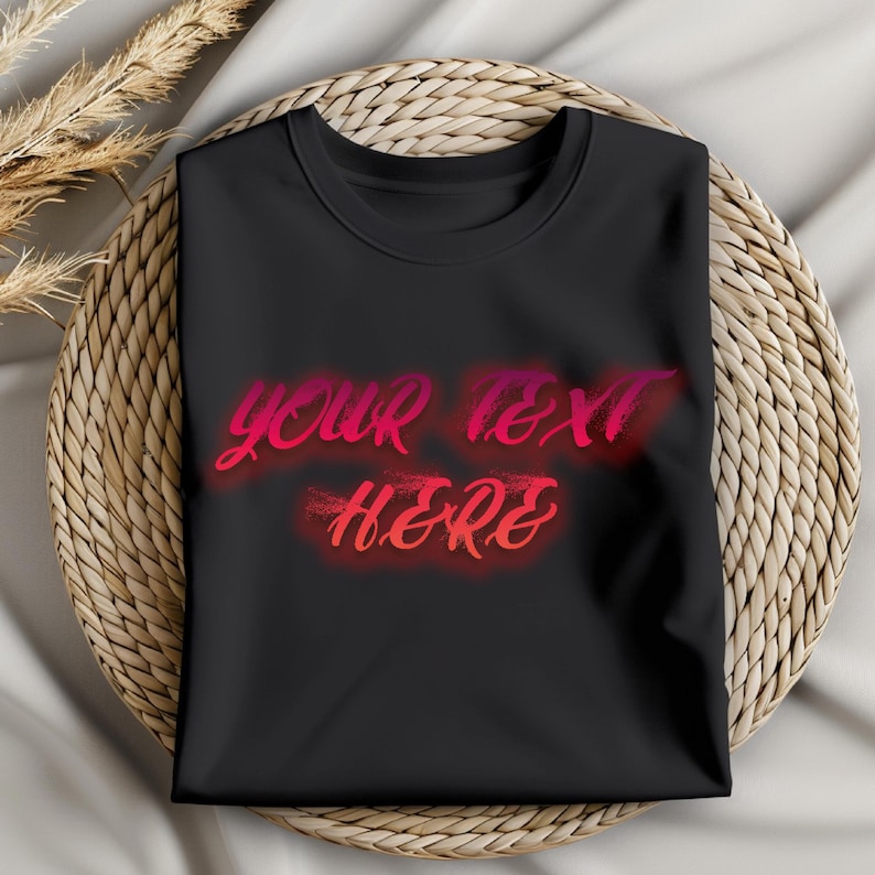 May include: A black t-shirt with the words "YOUR TEXT HERE" in a gradient red and pink font. The shirt is folded neatly and rests on a woven, circular placemat. Dried wheat stalks are visible in the upper left corner.