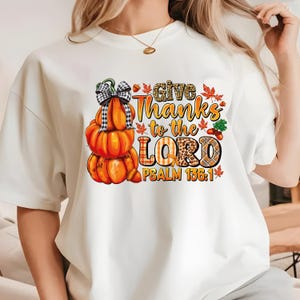 Peut inclure: T-shirt blanc avec un motif de pile de citrouilles avec un nœud et le texte "Give Thanks to the Lord Psalm 136:1".