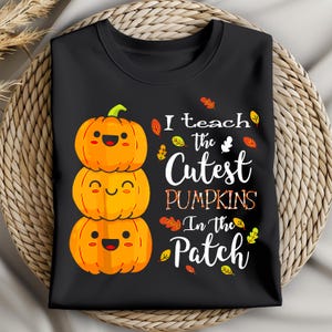 Peut inclure: T-shirt noir avec un motif de trois citrouilles oranges empilées, chacune avec un visage souriant. Le texte "I teach the Cutest Pumpkins In the Patch" est imprimé avec des feuilles. Un design d'automne saisonnier.