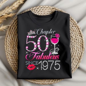 Puede incluir: Camiseta negra con un diseño gráfico que celebra un 50 cumpleaños. El diseño incluye el texto "Chapter 50 Fabulous Since 1975" con una tiara, una copa de vino con una bebida rosa y un par de labios rojos.