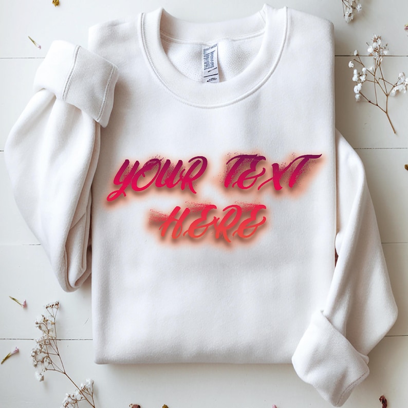 May include: A white sweatshirt with the text "YOUR TEXT HERE" in a gradient of pink and orange. The text has a spray-paint effect. The sleeves are cuffed. The sweatshirt is on a white wooden surface.