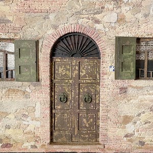 Può includere: Una facciata architettonica in miniatura con una porta marrone usurata con maniglie ad anello, affiancata da due finestre con persiane verdi. L'edificio ha un esterno in mattoni e pietra con un ingresso ad arco.