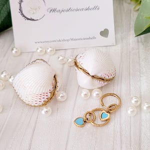 Può includere: Due portagioie a conchiglia bianchi con finiture dorate e un portachiavi dorato con ciondoli a forma di cuore turchese. Perle sparse e un biglietto da visita con "Majesticseashells".