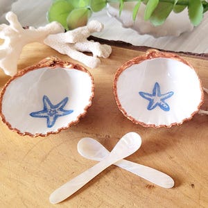 May include: Two white seashell dishes with a blue starfish design, and two small mother-of-pearl spoons. The dishes have a brown rim and are on a wooden surface. A tag reads "Salt and Pepper Dish".