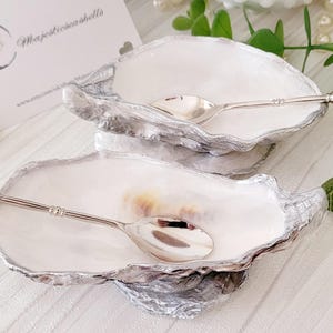 May include: Two oyster shell serving dishes with silver rims and spoons. Each shell has a white interior and a silver spoon with a crystal-like handle. A business card with the text "Majesticshells" is in the background.