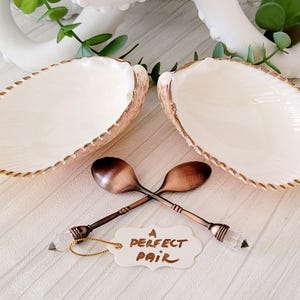 May include: Two white seashell bowls with gold trim and a pair of copper spoons with crystal handles. The spoons are crossed and the text "A Perfect Pair" is written on a white tag.