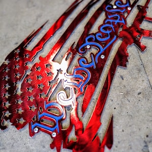 May include: A red, white, and blue metal American flag with the words "We the People" cut out in blue. The flag is distressed and has a torn look.