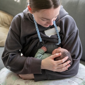 May include: A dark gray hooded sweatshirt with a light blue trim and a white patch. The sweatshirt is being worn while a baby in a light green onesie is being held.