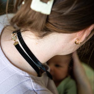 May include: A black velvet choker necklace with a gold charm and clear crystals. A gold hoop earring is visible. A white hair clip is in the hair. The necklace is worn around the neck.