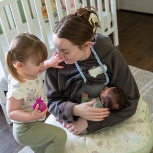 May include: A woman in a grey hoodie breastfeeds a baby, observed by a young child. The baby wears a green onesie. The woman sits on a patterned nursing pillow. A white cot is visible in the background.