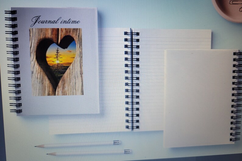 Puede incluir: Tres cuadernos de espiral con cubiertas blancas. La portada del cuaderno m&aacute;s a la izquierda tiene un recorte en forma de coraz&oacute;n con una imagen de un &aacute;rbol y una puesta de sol dentro. El texto "Journal intime" est&aacute; impreso en la portada.