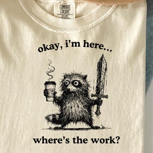 May include: Cream-colored t-shirt featuring a black and white illustration of a raccoon holding a coffee cup and a sword. The text "okay, I'm here... where's the work?" is printed below the raccoon.