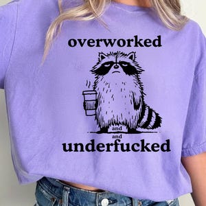 May include: A lilac t-shirt with a black and white illustration of a disgruntled raccoon holding a coffee cup. The text "overworked and underfucked" is printed above and below the raccoon graphic.