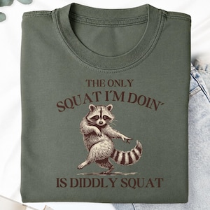 May include: An olive green crew neck t-shirt featuring a brown graphic illustration of a raccoon in a dancing pose. The text printed in brown above and below the raccoon reads, THE ONLY SQUAT I'M DOIN' IS DIDDLY SQUAT, in a bold, capitalized font.