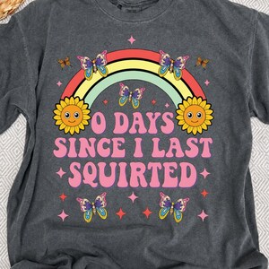 May include: Dark gray t-shirt with a rainbow, butterflies, and smiling sunflowers. The text reads "0 DAYS SINCE I LAST SQUIRTED" in pink and white. The shirt has a vintage, retro design.