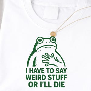 May include: White t-shirt featuring a green frog graphic with the text "I HAVE TO SAY WEIRD STUFF OR I'LL DIE". The frog is drawn in a simple, cartoon style. A gold necklace with a small pendant is also visible.