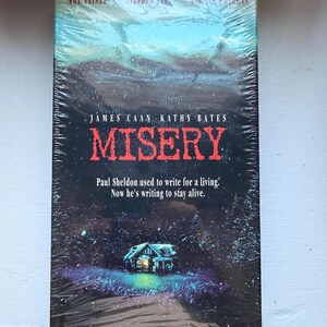 May include: A sealed VHS tape of the movie "Misery" featuring James Caan and Kathy Bates. The cover art depicts a snowy landscape with a house and the title in red. Text reads "Paul Sheldon used to write for a living. Now he's writing to stay alive."