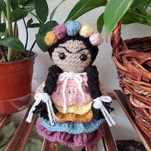May include: Crocheted doll with black hair, a colorful flower crown, and a multi-colored skirt. The doll is wearing a white crocheted top with a bow. The doll is sitting on a wooden surface.