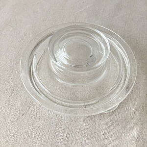 Puede incluir: Una tapa de vidrio transparente con diseño circular. La tapa presenta una parte central elevada con anillos concéntricos, rodeada por un borde plano más ancho. El vidrio es liso y transparente, adecuado para un tarro o recipiente. Diámetro aproximado de 10 cm.