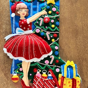 Può includere: Una calza natalizia in feltro con sfondo blu e un albero di Natale verde decorato con ornamenti colorati, bastoncini di zucchero e una stella gialla. Una figura con abito rosso e cappello da Babbo Natale appende una pallina, con regali.