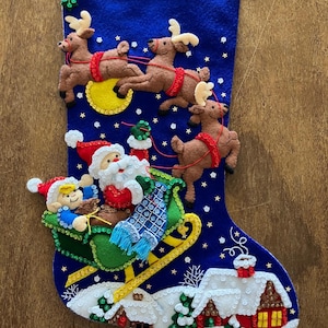 May include: A blue felt Christmas stocking featuring an appliqued scene of Santa Claus and an elf in a green sleigh pulled by three brown reindeer. The design includes a yellow moon, white stars, and a snowy village with houses at the bottom.