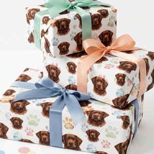 May include: Three rectangular gift boxes stacked vertically, wrapped in white paper featuring a repeating pattern of brown dog faces and pastel-colored paw prints. Each box is tied with a satin ribbon in mint green, peach, or dusty blue.