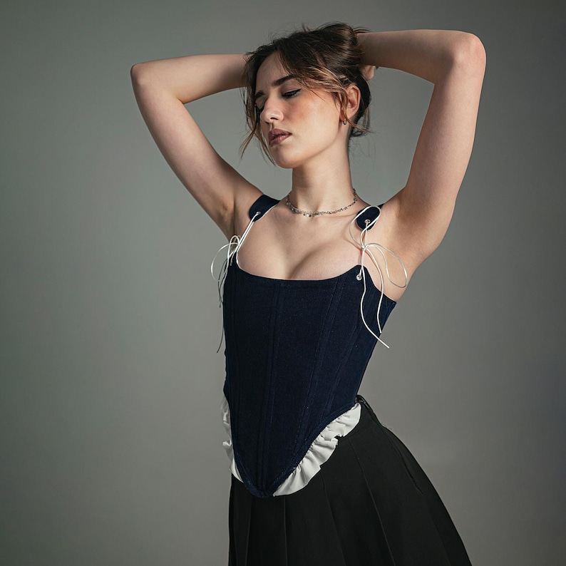 May include: A woman wearing a dark blue corset with white lace trim and a black skirt. She is standing with her arms raised above her head, and her eyes are closed.