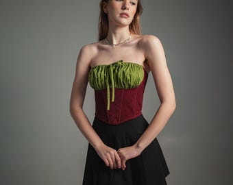 May include: A strapless corset top with a green gathered bust and a deep red bodice. The top has a bow detail at the center and is paired with a black pleated skirt. The outfit is a mix of textures and colors.