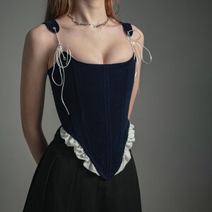 May include: A navy blue velvet corset with white lace trim and white ribbon straps. The corset has a boned structure and a low neckline.