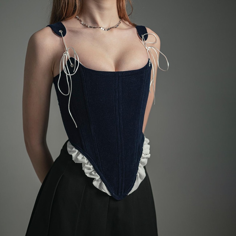 May include: A navy blue velvet corset with white lace trim and white ribbon straps. The corset has a boned structure and a low neckline.