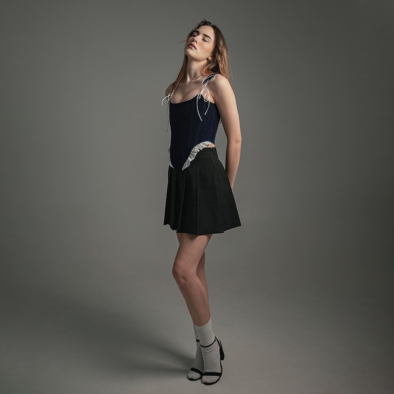 May include: A woman wearing a dark blue corset with white trim and a black pleated skirt. She is wearing white socks and black heels.