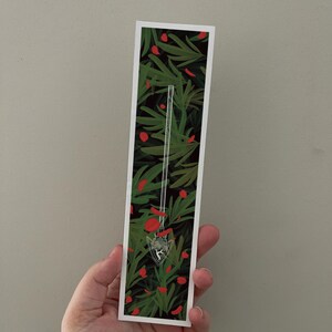 以下が含まれることがあります： 緑と赤の花柄と銀色の矢のデザインのしおりです。