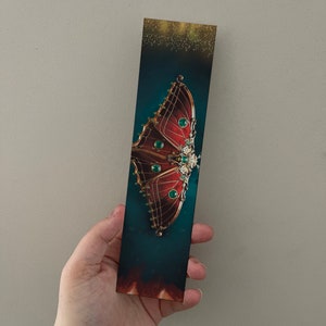 Puede incluir: Un marcapáginas con una ilustración detallada de una mariposa con detalles rojos, dorados y verdes sobre un fondo azul oscuro.
