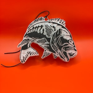 May include: A white car air freshener shaped like a fish with a black outline. The fish is hanging from a black string.