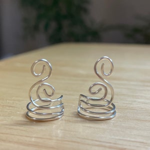 May include: A pair of silver ear cuffs with a swirling design. The cuffs are made of wire and have a delicate, feminine look.