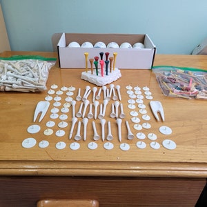 May include: A collection of white golf tees, white tee markers, and a set of colorful golf tees in a white box. The golf tees are arranged in a variety of patterns on a wooden surface.