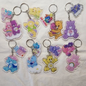May include: A collection of clear acrylic keychains featuring various Care Bears characters. Each keychain has a silver metal ring and chain. The bears are in different colors, including pink, purple, yellow, and blue, with unique designs and poses. Some keychains have the text "BE KIND".
