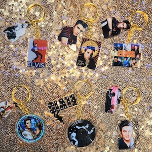 May include: A collection of gold keychains featuring images of Elvis Presley. The keychains have various designs, including portraits, album covers, and the name "ELVIS". The background is a sparkly gold.