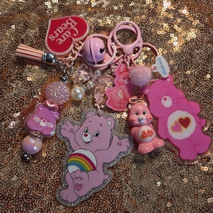 Puede incluir: Un llavero de Care Bears con múltiples dijes rosas, incluyendo un oso brillante con un arcoíris, una figura pequeña, una etiqueta en forma de corazón, una campana rosa, una borla y varias cuentas rosas y blancas en una anilla metálica.