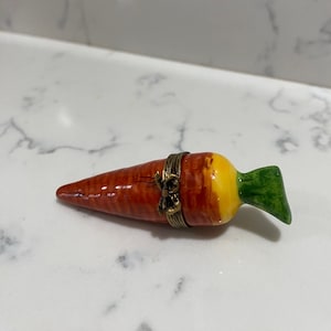 Könnte beinhalten: Eine kleine Keramik-Karotten-Schmuckschatulle mit einem goldfarbenen Scharnier und einem grünen Deckel. Die Karotte ist in Orange-, Gelb- und Grüntönen bemalt.