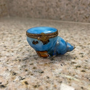 May include: A small blue porcelain trinket box shaped like a seashell with gold accents and a seashell design. The box has a hinged lid with a gold trim.