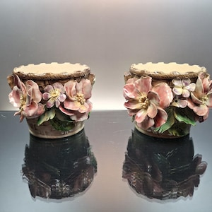 May include: A pair of ceramic planters featuring a brown woven basket texture and decorative three-dimensional pink flowers with green leaves. The planters have a scalloped top edge and a cylindrical shape, showcasing detailed floral craftsmanship.