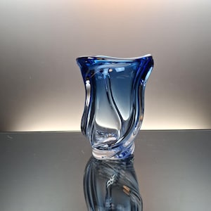 May include: A hand-blown glass vase featuring a deep cobalt blue hue that transitions into a clear, transparent base. The vessel has a fluid, organic shape with undulating, wave-like vertical ridges and a wide, asymmetrical, flared rim.