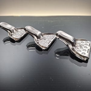 May include: Three stainless steel citrus juicers are arranged in a row. Each juicer has a handle and a perforated, triangular pressing surface. The juicers are on a reflective, dark surface.