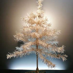 Könnte beinhalten: Ein kleiner, künstlicher Weihnachtsbaum mit metallisch-lamettaartigem Aussehen. Der Baum ist weiß mit einem goldfarbenen Stamm und Sockel. Die Zweige sind ausgebreitet, und der Baum steht auf einem goldfarbenen, sternförmigen Sockel.