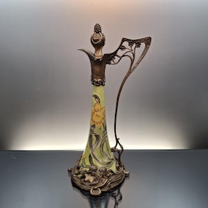 May include: A tall, slender glass decanter with a pale green body featuring a painted yellow daffodil and dark green leaves. The decanter has a bronze-toned metal base with sculpted figures, a matching metal neck, and an ornate, sweeping handle.